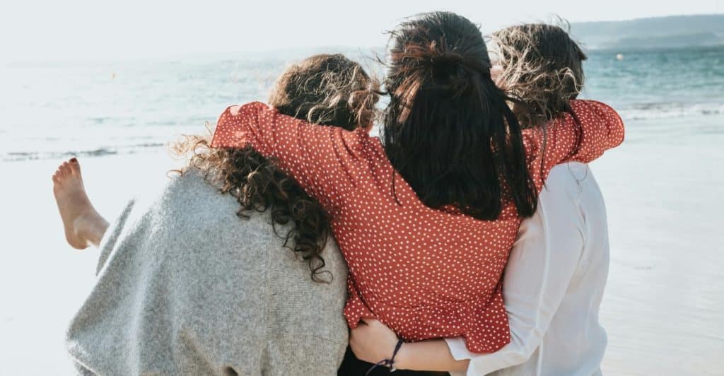 Three women, seen from behind, embracing on a beach with the ocean in the background.