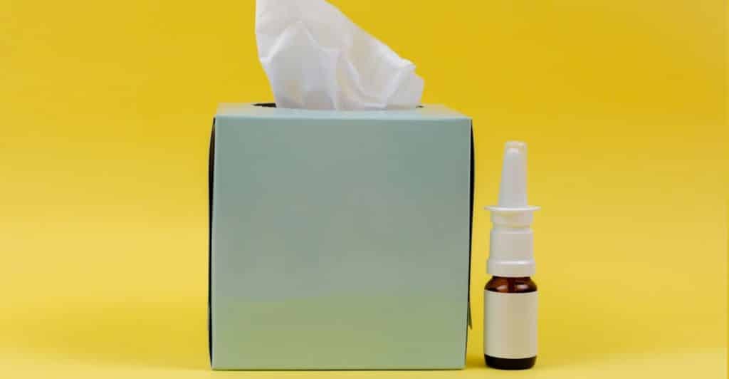A light blue box of tissues next to a nasal spray bottle on a bright yellow background.