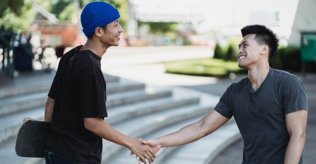 Two men of Asian descent shaking hands outdoors, smiling at each other.