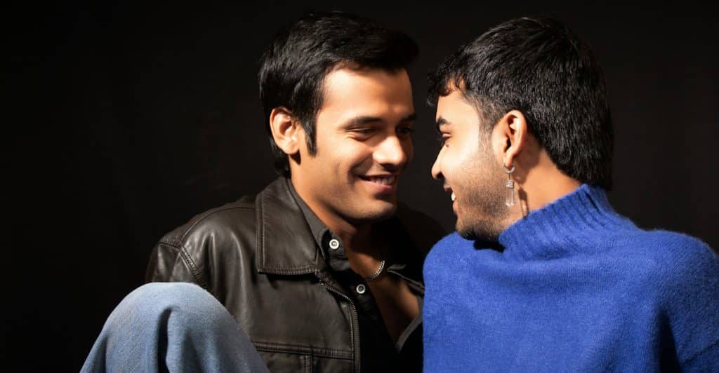 Two men, one in a black leather jacket and the other in a blue turtleneck, looking at each other and smiling with a dark background.
