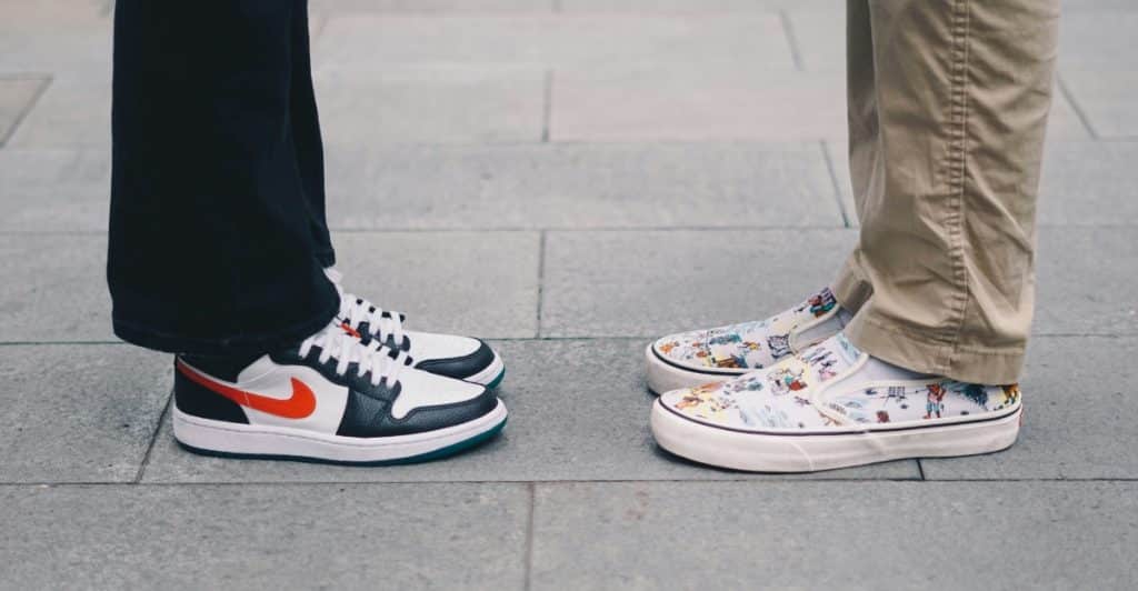 The lower legs and feet of two people facing each other, one wearing black and white Nike sneakers and the other wearing white Vans slip-ons with colorful drawings.