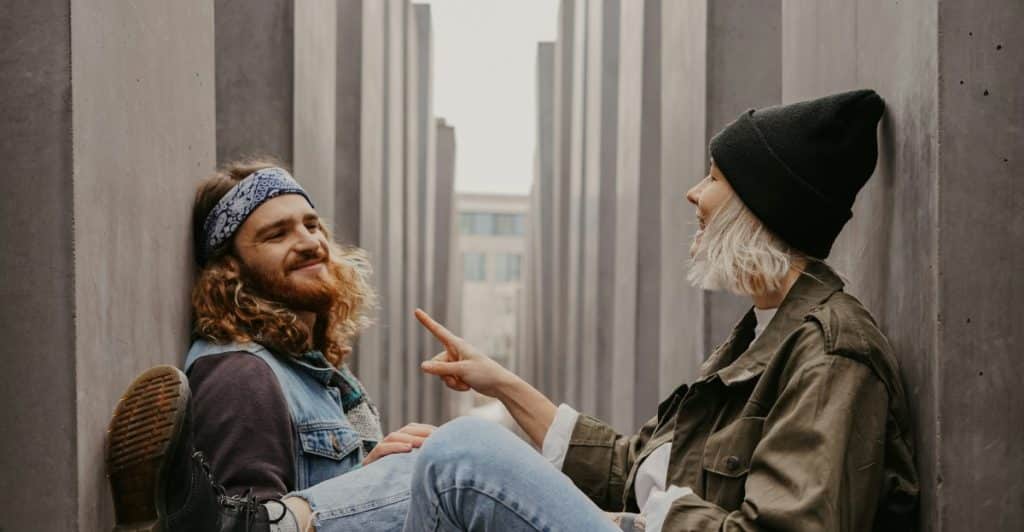 A man with long, curly red hair and a bandana sitting against a concrete pillar, talking and smiling at a woman with short blonde hair and a black beanie, who is also sitting against a pillar.