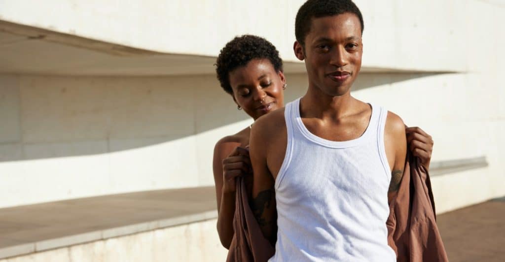 A woman standing behind a man, pulling a brown shirt off his shoulders, while the man, wearing a white tank top, looks forward.