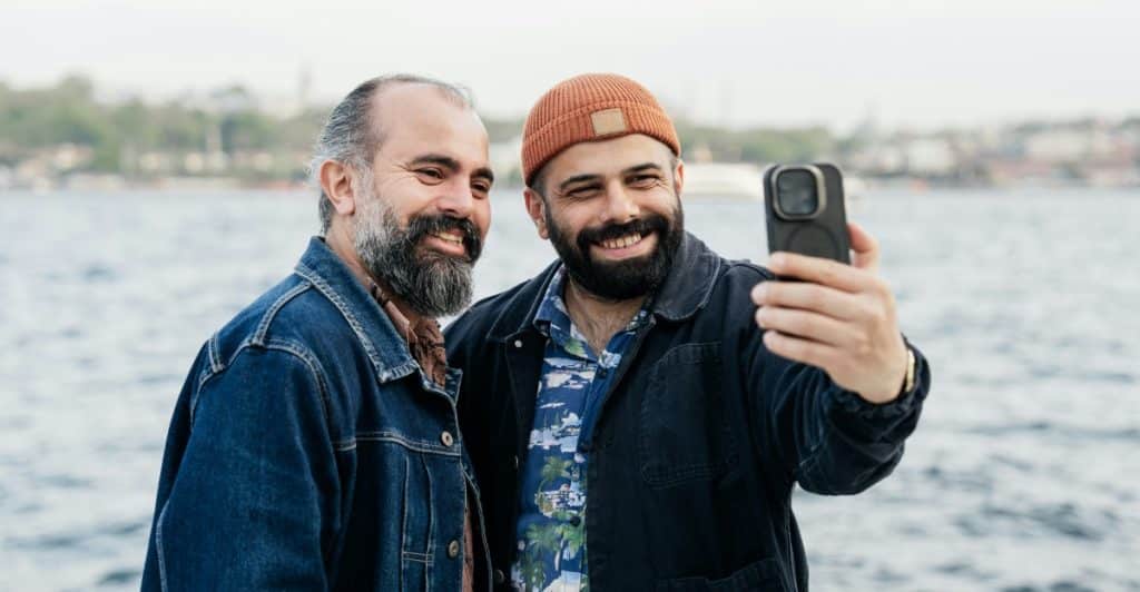 Two bearded men smiling and taking a selfie together outdoors near a body of water.