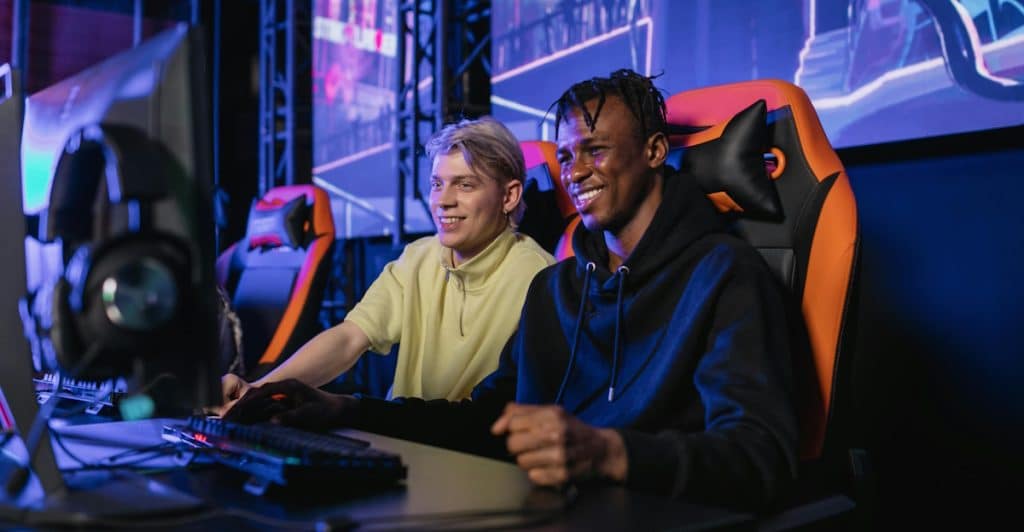 Two men are seated at gaming stations, both smiling and engaged in playing video games in a dimly lit room with vibrant blue and pink neon lights in the background.