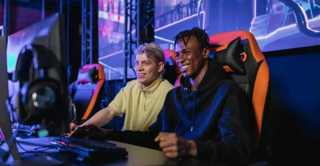 Two young men, one with blonde hair and one with dreadlocks, sitting at computer desks with keyboards and monitors, smiling and looking at each other, with colorful screens in the background.