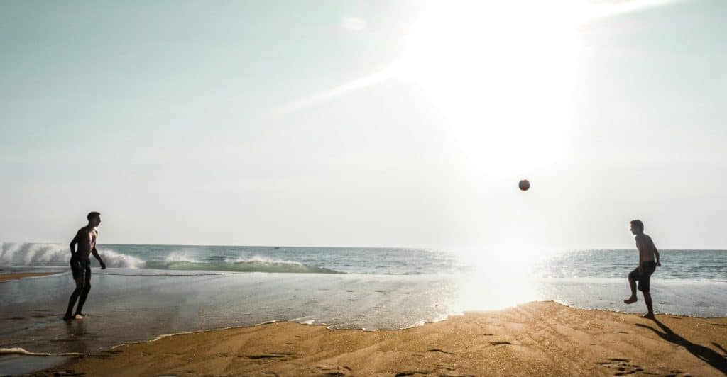 Two men playing soccer on a beach near the ocean with a ball in the air.
