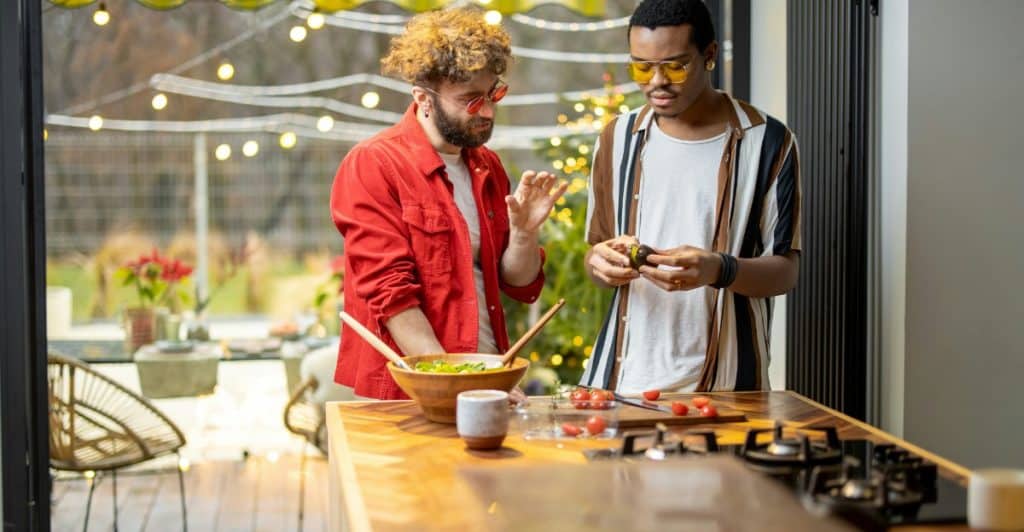 Two men, one with a beard and red beanie, and the other with sunglasses, preparing food on a counter outdoors