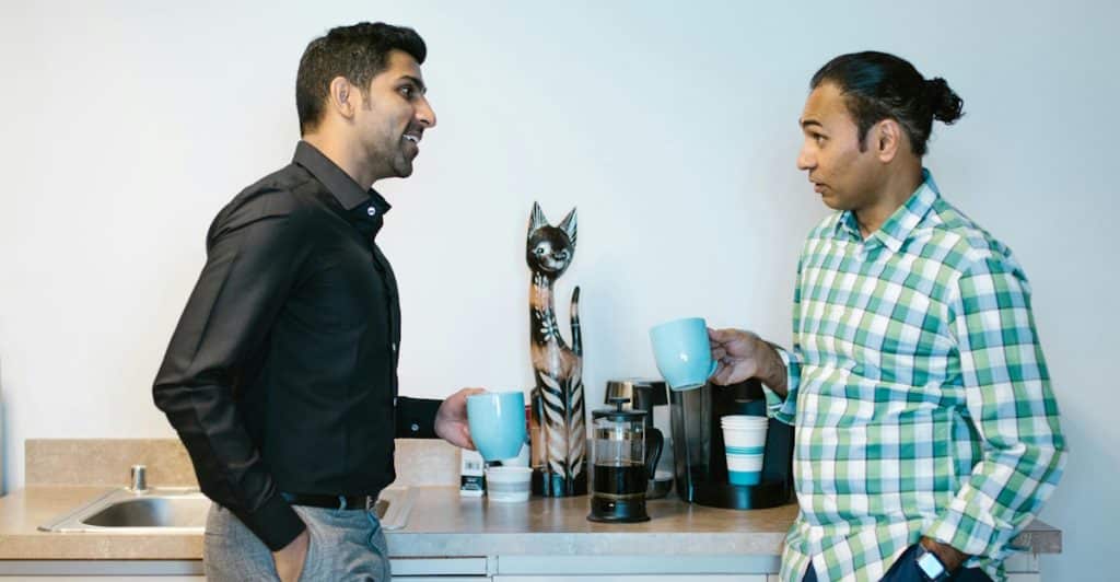Two men are standing in a light-colored room with a coffee machine on a counter behind them; the man on the left is wearing a dark shirt and talking while holding a blue mug, and the man on the right is listening while holding a similar blue mug.