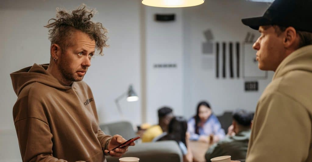 Two men are standing and facing each other, engaged in conversation; the man on the left has curly hair and is holding a phone and a white cup, while the man on the right is wearing a black cap and holding a similar cup, with a laptop between them.