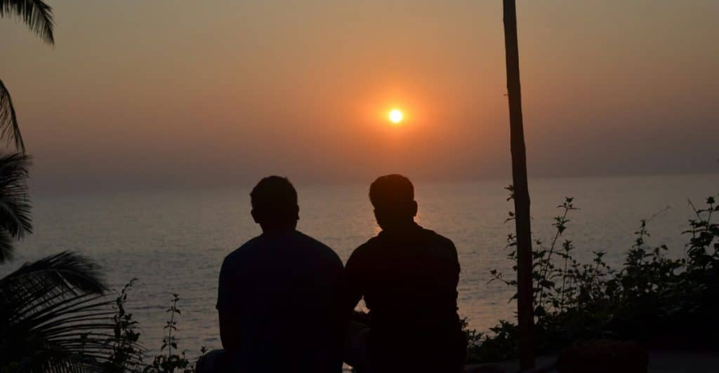 Two men seen from behind, sitting on the ground and looking out at a sunset over a body of water.