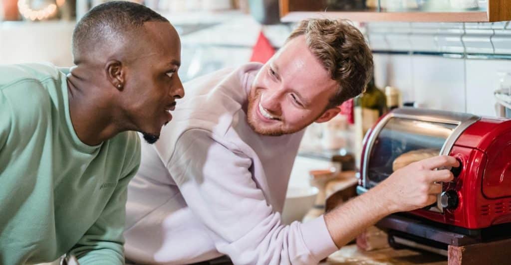 A man with a fade haircut and a light green long-sleeve shirt looking at another man with a beard and light-colored hair, who is smiling and adjusting a red toaster oven on a wooden counter.
