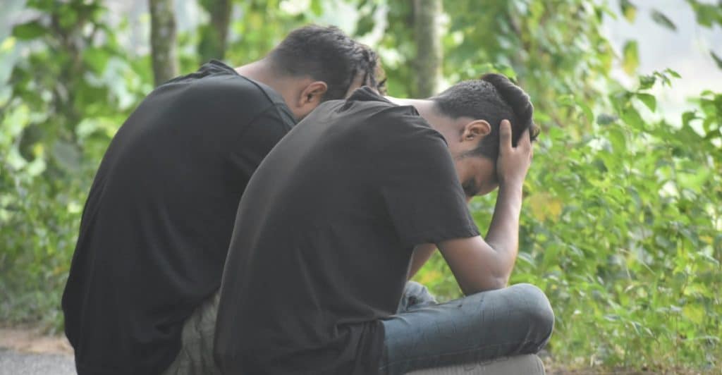 Two men sitting on a paved surface with their backs to the viewer and hands on their heads.
