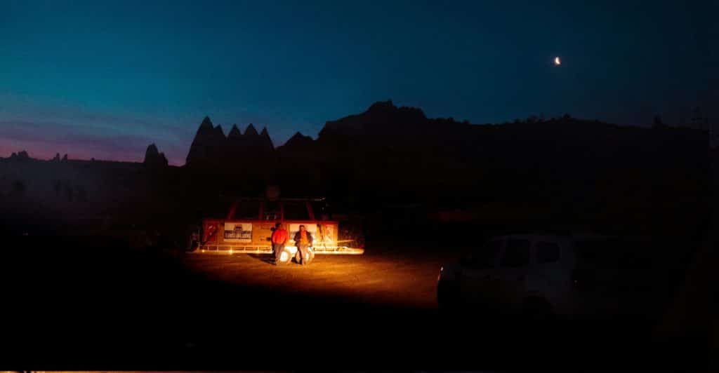 Two people standing at night near a brightly lit structure with mountains in the background and a crescent moon in the sky.