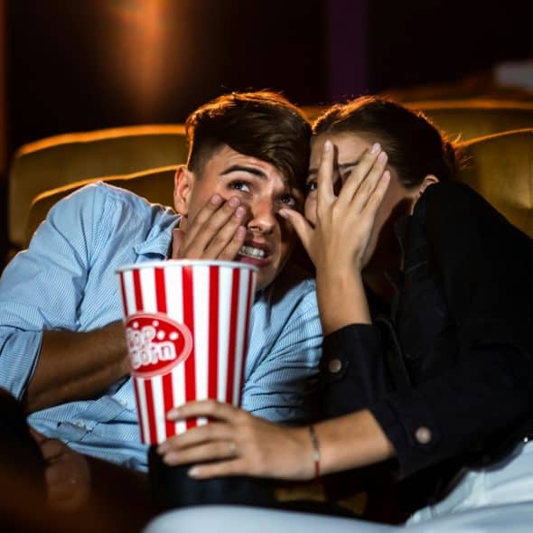 A man and a woman covering their faces with their hands while sitting in a movie theater with a bucket of popcorn.
