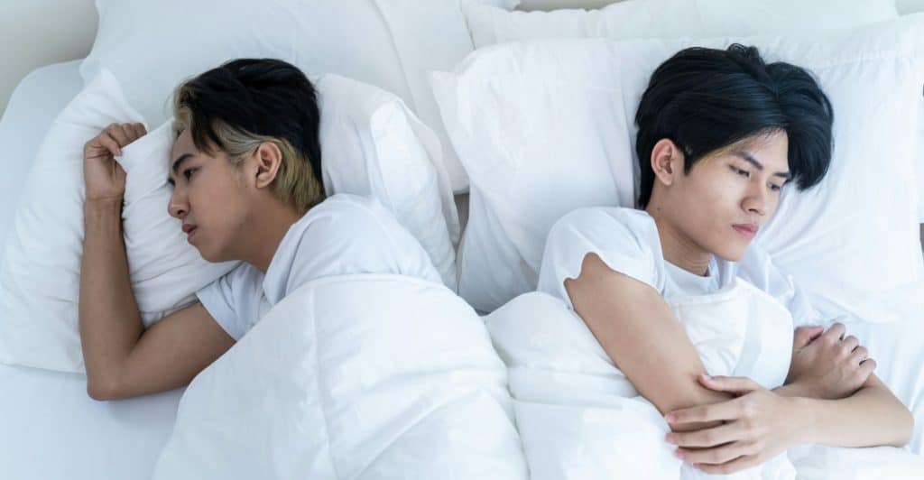 Two people with dark hair lying in bed facing away from each other, both appearing sad or distant.
