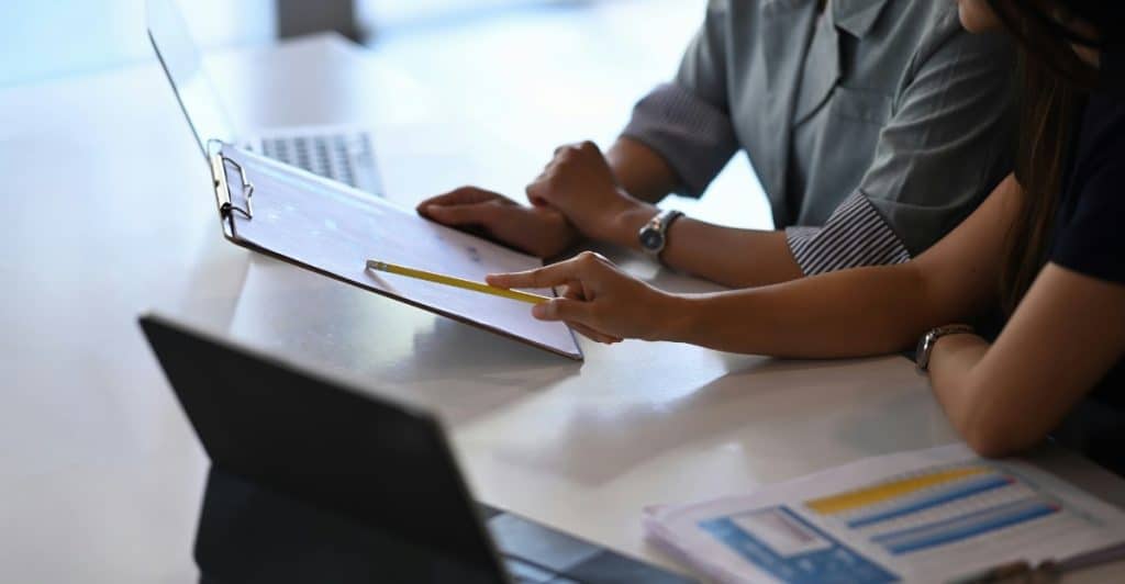Two people at a desk, one holding a pencil and pointing at a document on a clipboard, with a laptop and papers nearby.