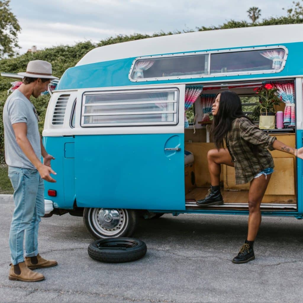 A person on the right stepping out of a vintage blue and white van and gesturing with their hands while facing another person on the left who is wearing a hat and light-colored clothes
