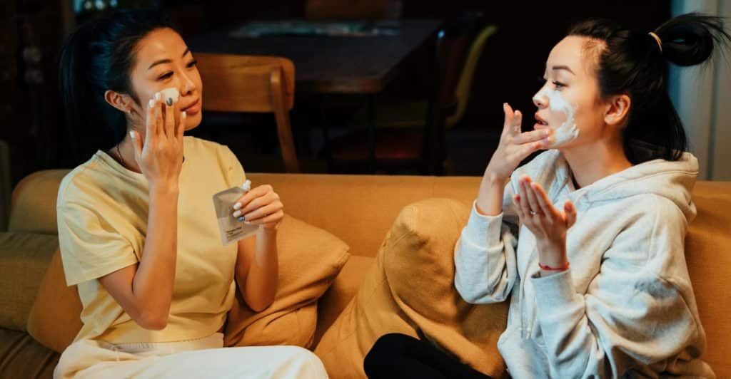 Two women sitting on a couch, applying skincare products to their faces.