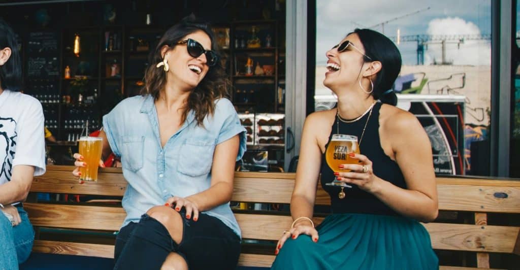 Two women wearing sunglasses, laughing and holding drinks at an outdoor cafe.