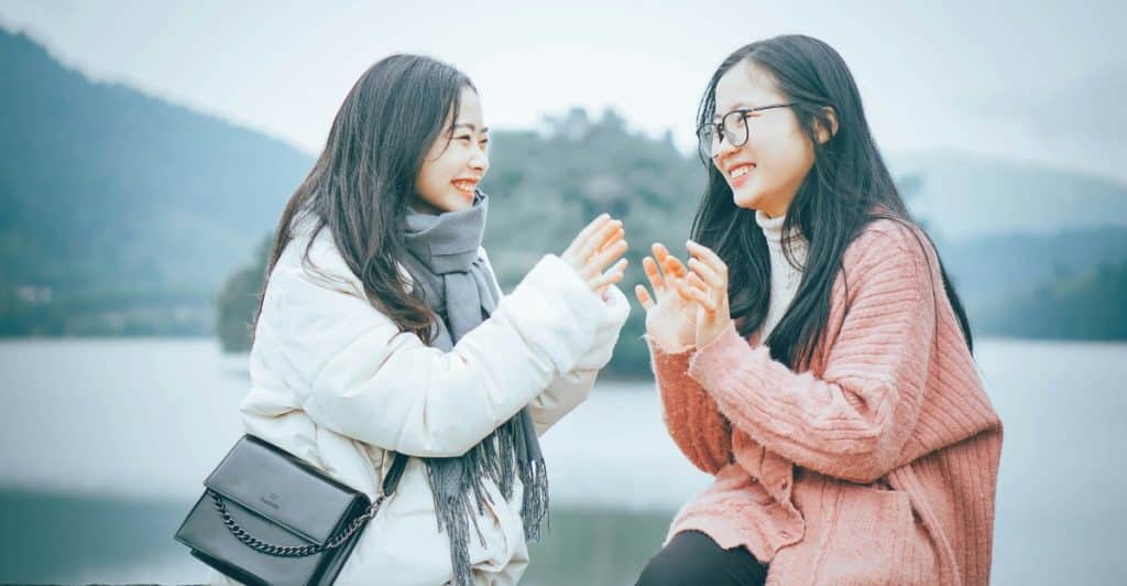 Two young women, of Asian descent, bundled in winter clothing and laughing while sitting outdoors with a body of water behind them