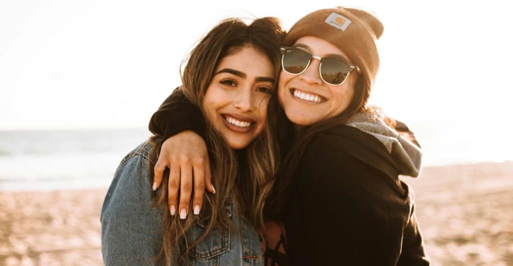 Two women smiling and hugging on a beach with the ocean in the background