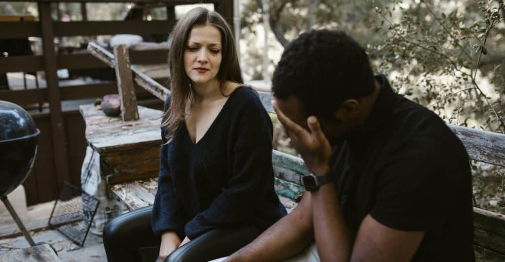 A woman looking at a man who is sitting with his head in his hands on a wooden bench outdoors.