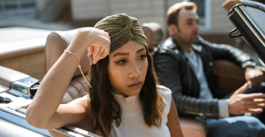 A woman wearing a turban and white top sitting in the passenger seat of a convertible car, looking away with an upset expression, with a man in the driver's seat.