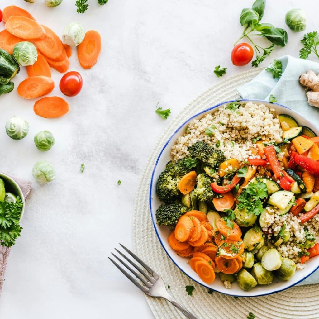 A vibrant flat-lay photograph showcasing a vegetable salad in a white bowl, surrounded by fresh carrots, zucchini, tomatoes, and Brussels sprouts on a light background with a fork and a napkin.