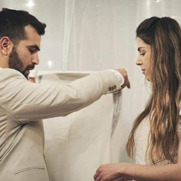 A groom in a light-colored suit gently adjusting the long white veil of his bride, who is wearing a white lace wedding gown and looking down.