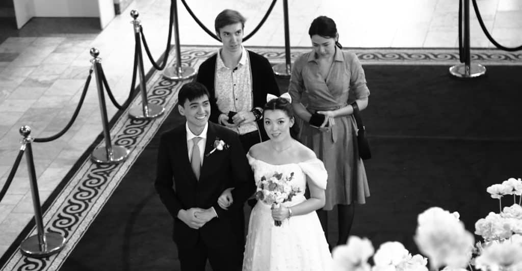 A black and white photo of a bride and groom standing on a patterned carpet, with two other people standing behind them.