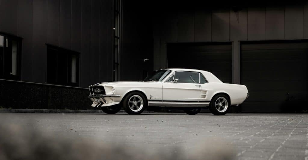 A white classic Ford Mustang with black racing stripes parked on an asphalt lot in front of a dark building.