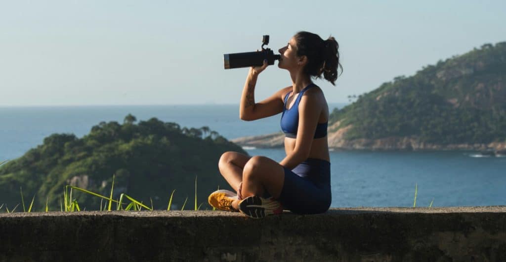 A woman sitting outdoors, drinking from a water bottle with a scenic view in the background