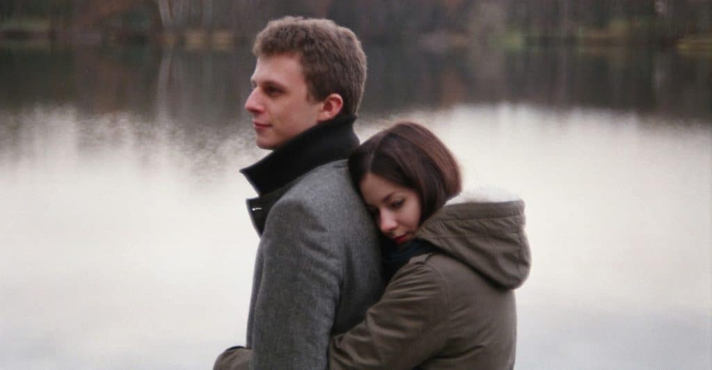 A woman with dark hair hugging a man from behind by a lake, resting her head on his back.