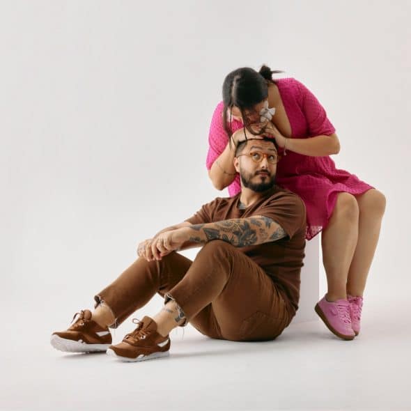 A man with a beard and tattoos is sitting on the floor while a woman in a pink dress is sitting on a white block behind him, gently kissing his head.