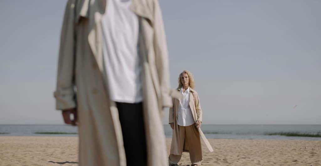 A woman in a beige coat looking towards a man in the foreground on a sandy beach, with a calm body of water and clear sky in the background.