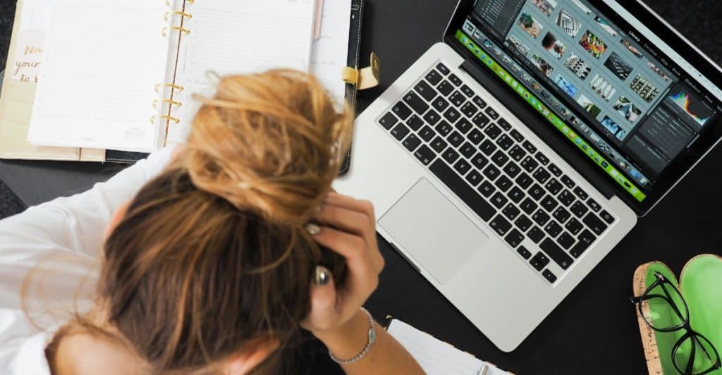 A woman with her head in her hands, looking stressed at a desk with a laptop and notebook.