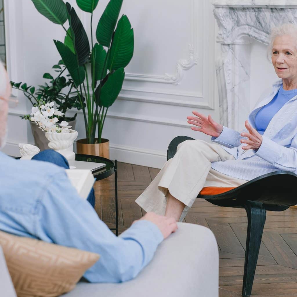 A woman with gray hair wearing a light blue top and white shirt talking animatedly to a man while sitting on a patterned chair in a well-decorated living room.