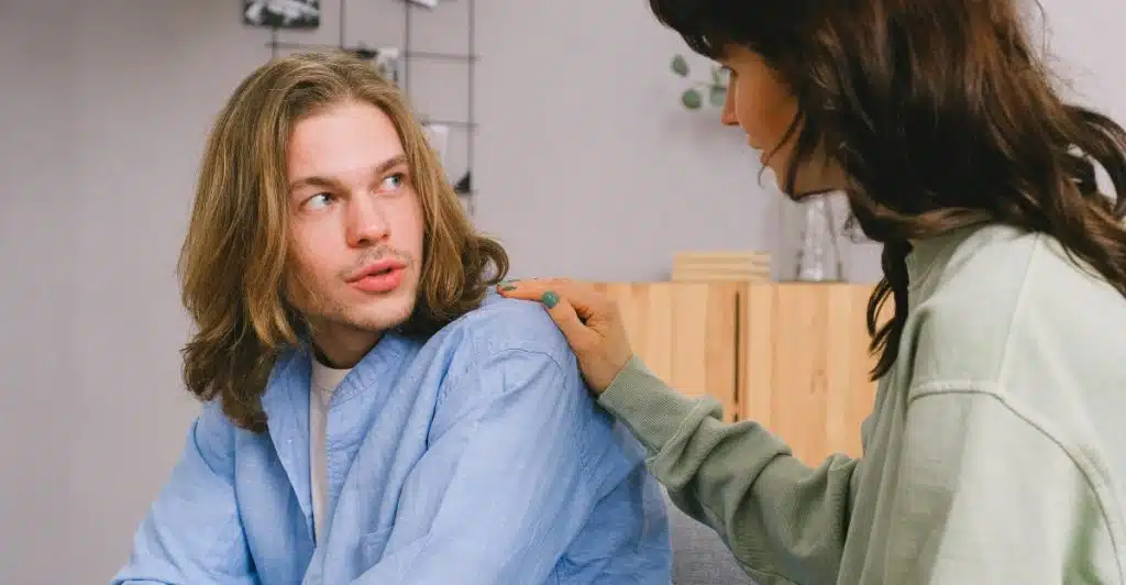 A woman with long dark hair, wearing a light green sweatshirt, gently places her hand on the shoulder of a man with long blond hair in a blue shirt, as they sit and converse in a softly lit room.