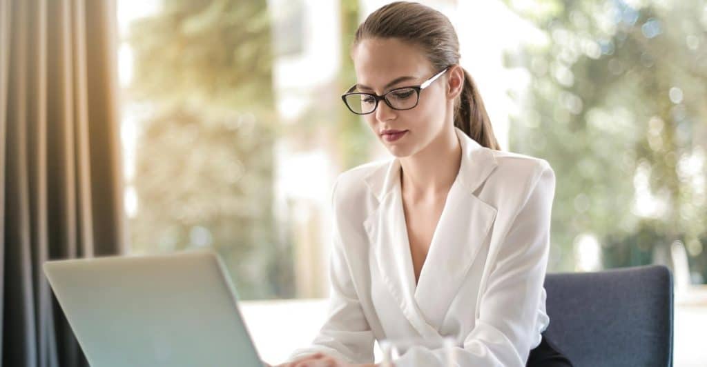 A focused female entrepreneur wearing glasses and a white blazer is seated at a table, diligently typing on a silver laptop, with a glass of water and a notebook nearby in a bright office setting.