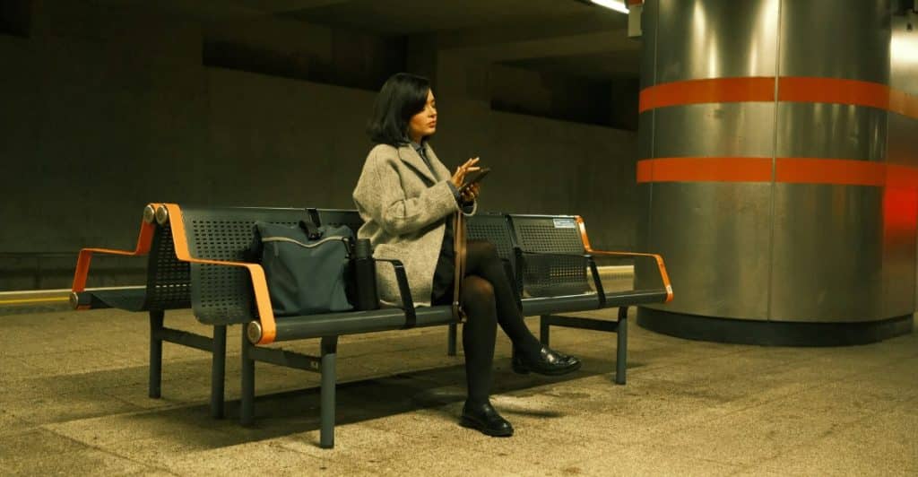 A woman with dark hair wearing a long coat and black tights sitting on a public bench at a subway station, looking at her phone.