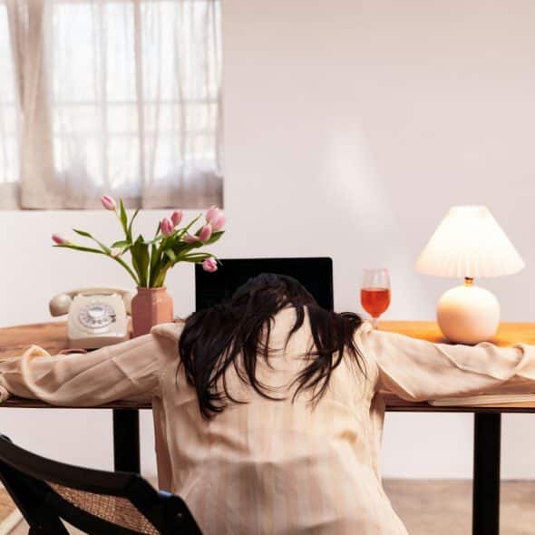 A woman with her head down on a desk, appearing exhausted, with a laptop, lamp, and flowers nearby.