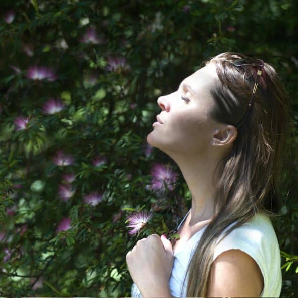 A woman with her eyes closed and head tilted up, standing in front of green foliage with some purple flowers, suggesting a moment of peace or meditation.