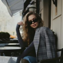 A woman in a black top, plaid blazer, and black sunglasses is seated outdoors at a cafe table.