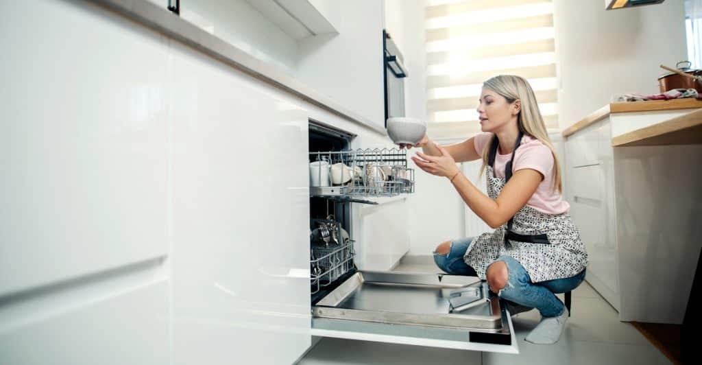 A woman crouching and loading a white bowl into a smart dishwasher.