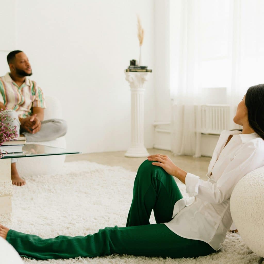 A woman sitting on a white rug with green pants and a white shirt, looking over her shoulder towards a man sitting on a white couch in a bright, minimalist room.