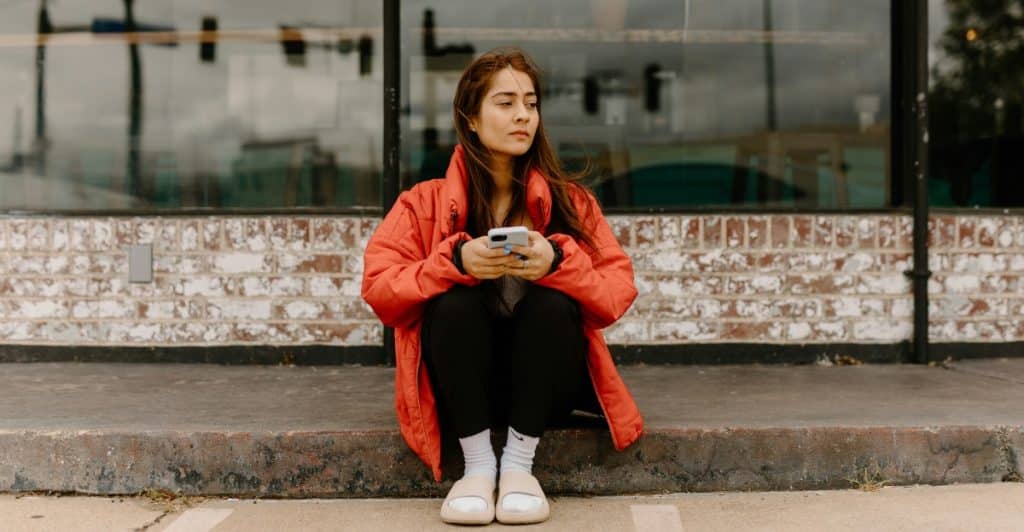 A woman with long brown hair, possibly of South Asian descent, sitting on a curb and looking contemplative while holding her phone.