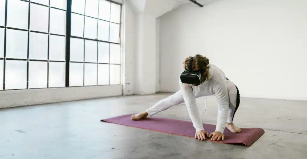 A woman wearing a VR headset and activewear stretching on a yoga mat in a large, bright, empty room with industrial windows.