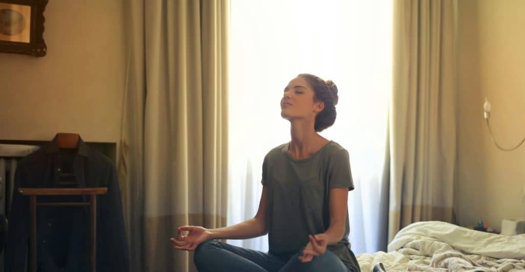 A woman sitting cross-legged on a bed in a bedroom, with her eyes closed and hands resting on her knees, in a meditative pose.