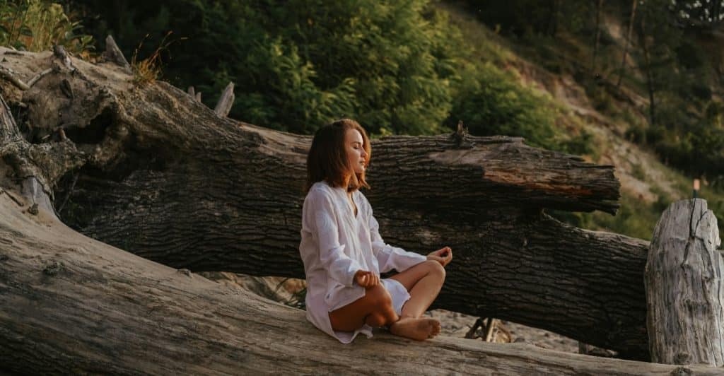 A woman sitting cross-legged on large tree roots outdoors, with her eyes closed, appearing to meditate.